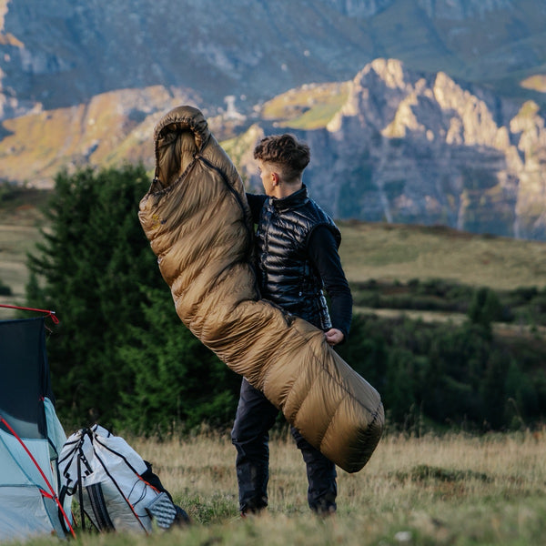 Person carrying X-Lite 400 sleeping bag in a scenic outdoor setting with mountains and trees.
